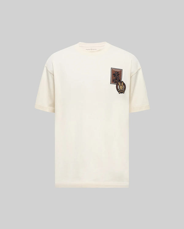 The Psycho Bunny MENS CAEL GRAPHIC TEE (MT0200181) is a regular fit off-white cotton tee featuring a small brown and black horse graphic patch on the upper left chest, shown on a light gray background.