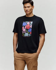 A man with short dark hair wears the Psycho Bunny MENS ROWEN OVERSIZED TEE (MT0200194), a relaxed fit black T-shirt featuring a colorful floral graphic and "Psycho Bunny" text, paired with light brown cargo pants against a plain light background.