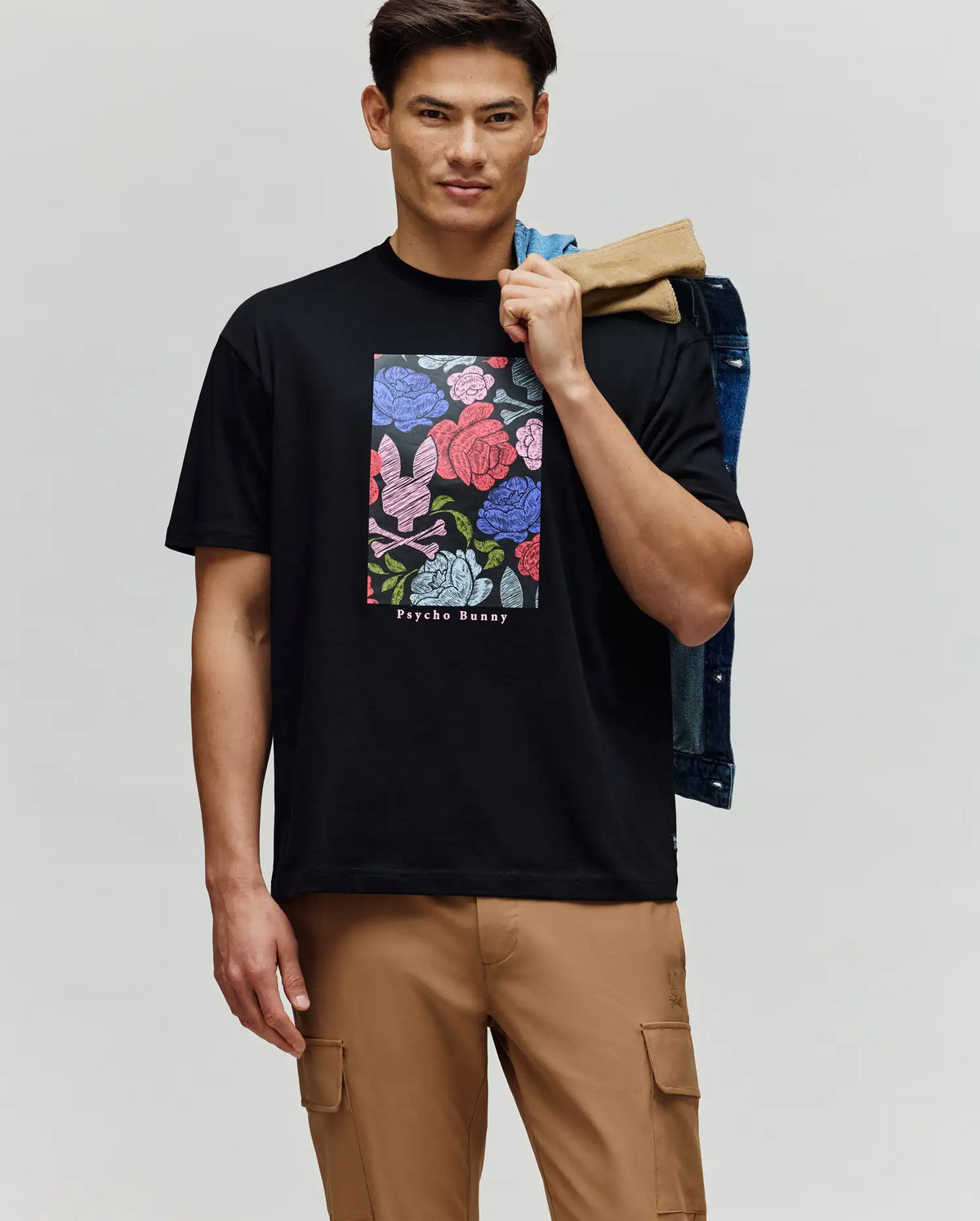 A man wears the Psycho Bunny MENS ROWEN OVERSIZED TEE - MT0200194, a relaxed black T-shirt with a colorful floral graphic and logo text. He holds a denim jacket over his shoulder, pairs it with tan cargo pants, and stands against a plain background.
