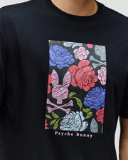 A person wears the Psycho Bunny MENS ROWEN OVERSIZED TEE (MT0200194), a relaxed black tee with a vibrant floral graphic—pink and blue roses with a bunny skull and crossbones—and "Psycho Bunny" below. Made from soft Pima cotton.