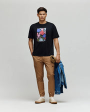 A man stands against a plain background wearing the Psycho Bunny MENS ROWEN OVERSIZED TEE (MT0200194), a relaxed fit black Pima cotton t-shirt with a colorful floral graphic, paired with tan cargo pants and white sneakers. He holds a denim jacket and brown cap.