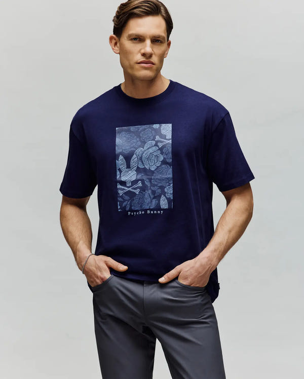 A man stands with his hands in his pockets, wearing the Psycho Bunny MENS ROWEN OVERSIZED TEE - MT0200194 in navy blue with a light blue floral and bunny chest graphic, paired with dark gray pants and looking at the camera.