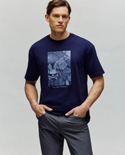 A man stands with his hands in his pockets, wearing the Psycho Bunny MENS ROWEN OVERSIZED TEE - MT0200194 in navy blue with a light blue floral and bunny chest graphic, paired with dark gray pants and looking at the camera.