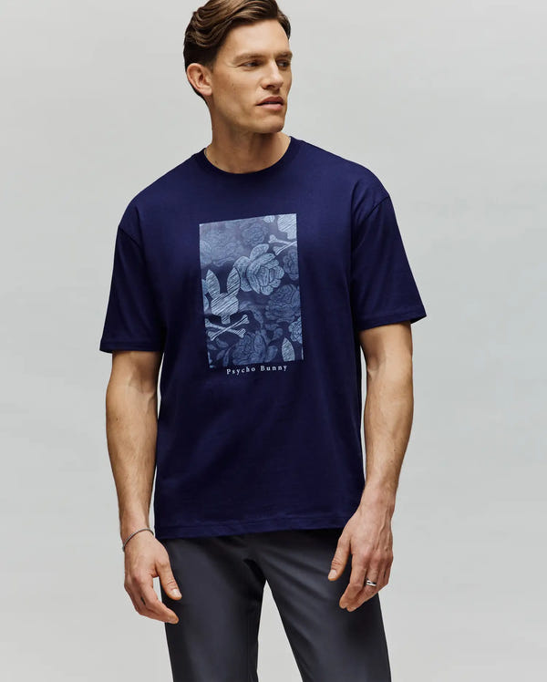 A man with short brown hair, looking to the side, wears the Psycho Bunny MENS ROWEN OVERSIZED TEE (MT0200194) in navy blue, featuring a relaxed fit and a rectangular floral "Psychic Beauty" graphic, against a plain light gray background.