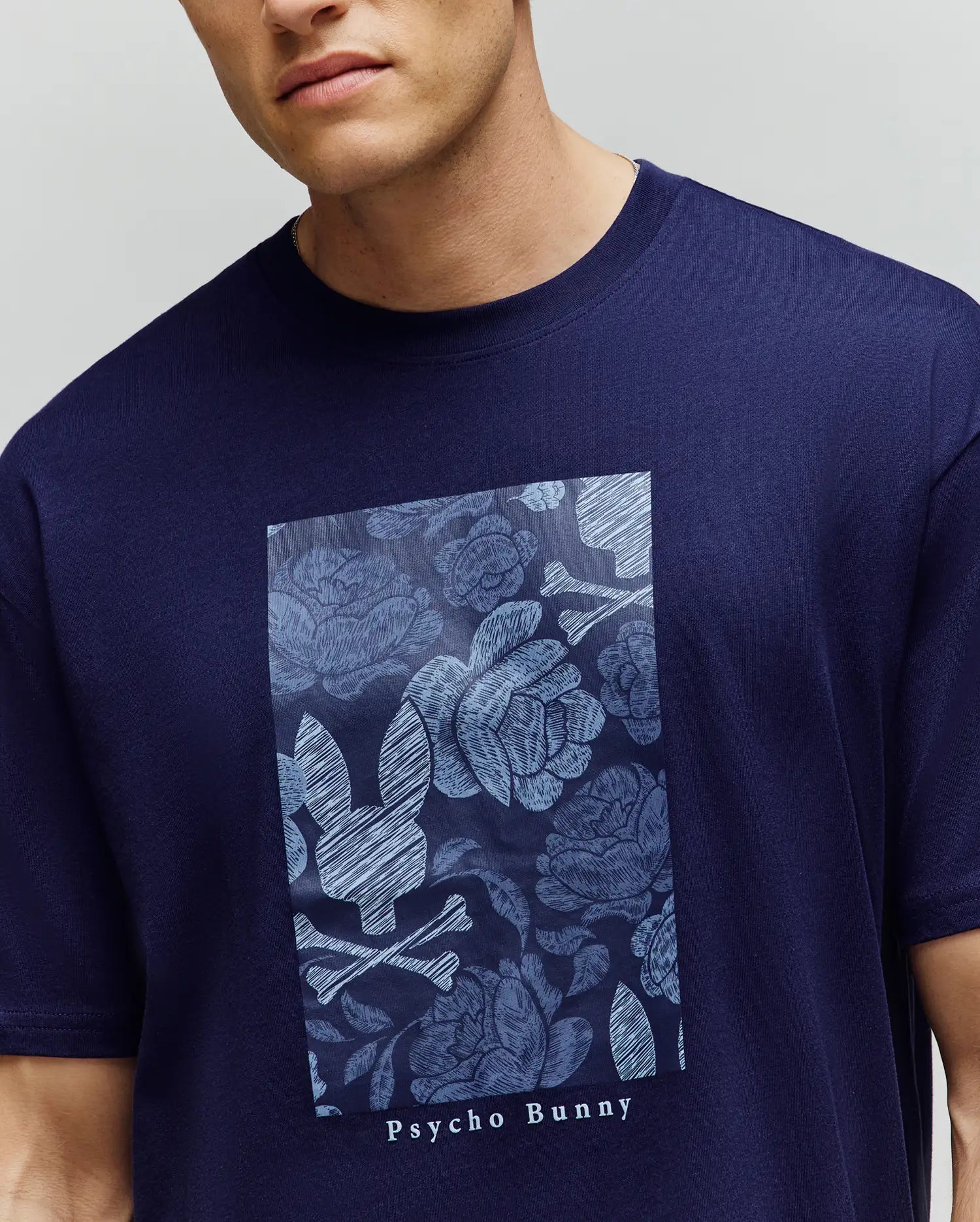 A man wearing the Psycho Bunny MENS ROWEN OVERSIZED TEE (MT0200194), a relaxed fit navy Pima cotton shirt with a bold floral graphic and large bunny skull logo on the front.
