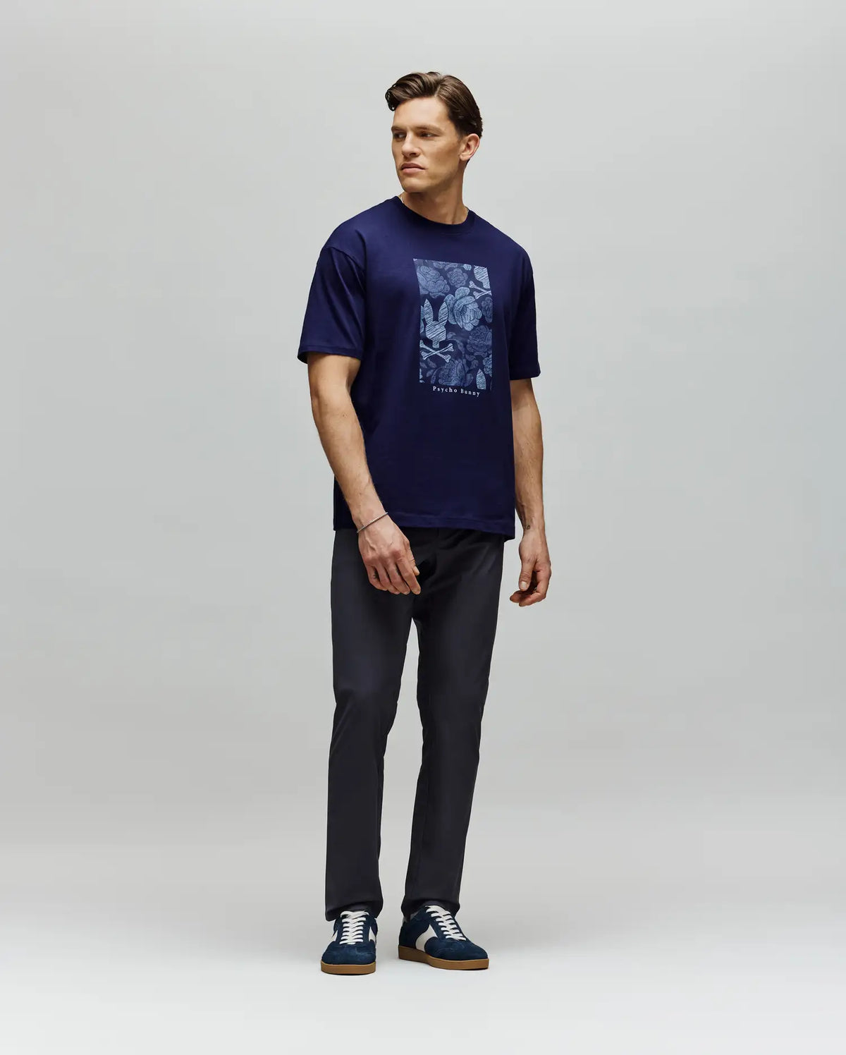 A man stands against a gray background wearing the Psycho Bunny MENS ROWEN OVERSIZED TEE (MT0200194) in navy with a floral graphic, paired with dark pants and navy sneakers. He looks left, one arm relaxed by his side and the other slightly bent.