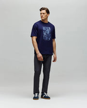 A man stands against a gray background wearing the Psycho Bunny MENS ROWEN OVERSIZED TEE (MT0200194) in navy with a floral graphic, paired with dark pants and navy sneakers. He looks left, one arm relaxed by his side and the other slightly bent.