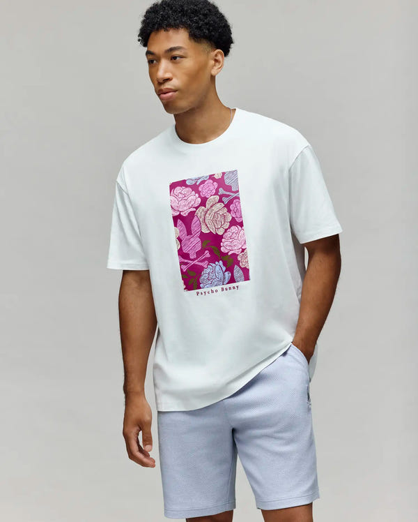 A man stands against a plain background wearing the Psycho Bunny MENS ROWEN OVERSIZED TEE - MT0200194, a white Pima cotton t-shirt with a vibrant floral graphic and "Psycho Bunny" text, paired with light gray shorts.