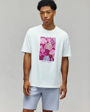 A man wears the Psycho Bunny MENS ROWEN OVERSIZED TEE (MT0200194), a white relaxed fit tee featuring a vibrant floral graphic and "Psycho Bunny" text, paired with light gray shorts against a plain light background.