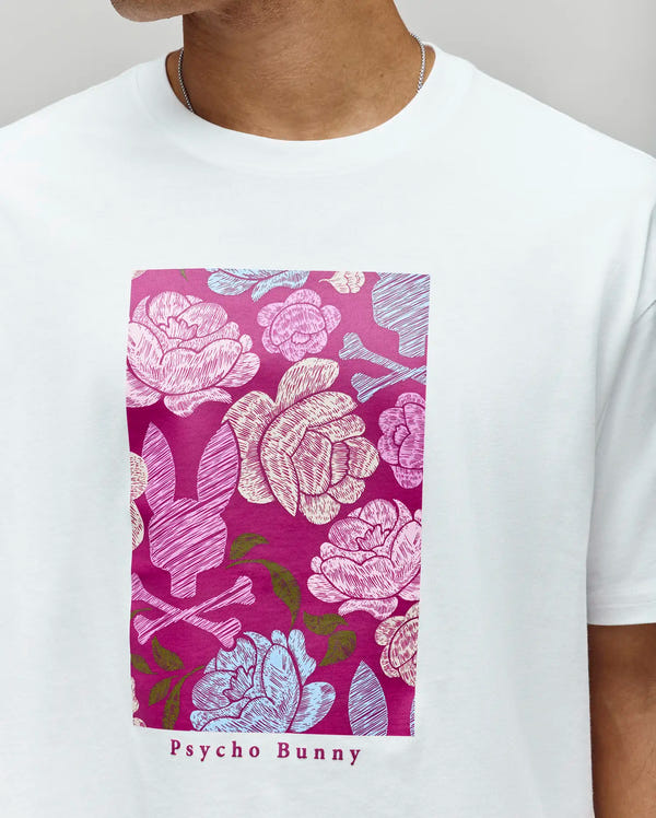 A person wears the Psycho Bunny MENS ROWEN OVERSIZED TEE (MT0200194), a relaxed fit Pima cotton shirt with a rectangular floral graphic—pink, purple, and blue flowers with bunny silhouettes—and "Psycho Bunny" text below.