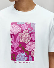 A person wears the Psycho Bunny MENS ROWEN OVERSIZED TEE (MT0200194), a relaxed fit Pima cotton shirt with a rectangular floral graphic—pink, purple, and blue flowers with bunny silhouettes—and "Psycho Bunny" text below.