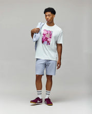 A man poses against a plain background wearing the Psycho Bunny MENS ROWEN OVERSIZED TEE (MT0200194) with a pink floral graphic, light blue shorts, maroon sneakers, white socks with black stripes, and a light blue sweatshirt over his shoulder.