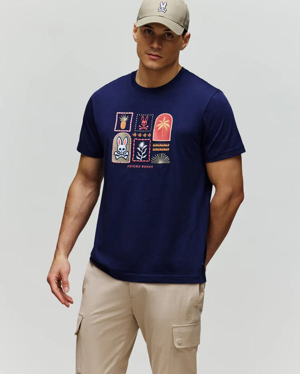 A man stands against a light gray background wearing the Psycho Bunny MENS SUNDEW GRAPHIC TEE (MT0200195) in soft Pima cotton with colorful squares, a beige cap, and light beige cargo shorts.