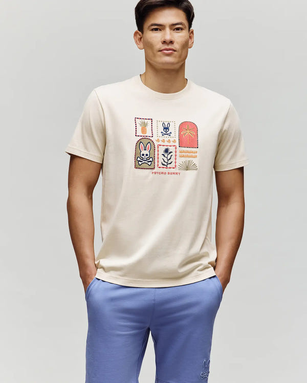 A man in the Psycho Bunny MENS SUNDEW GRAPHIC TEE (MT0200195), a cream Pima cotton shirt with colorful patches and branding, pairs it with light blue shorts and poses hands-in-pocket against a plain backdrop, embodying ocean view style.