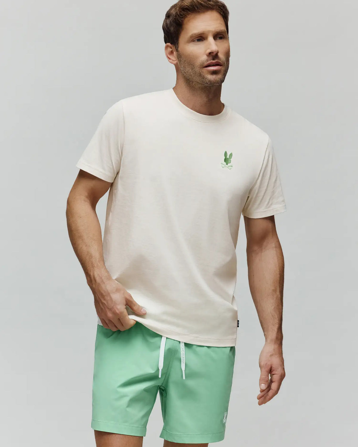 Mens Aiden Back Graphic Tee, a classic fit off-white jersey t-shirt featuring a small, vibrant green embroidered Psycho Bunny skull logo on the left chest.