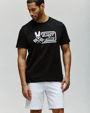 A man in a Psycho Bunny MENS RACING GRAPHIC TEE (MT0200243) with crossed tennis rackets and white shorts stands against a plain light background, glancing down to his left for a relaxed, stylish look.