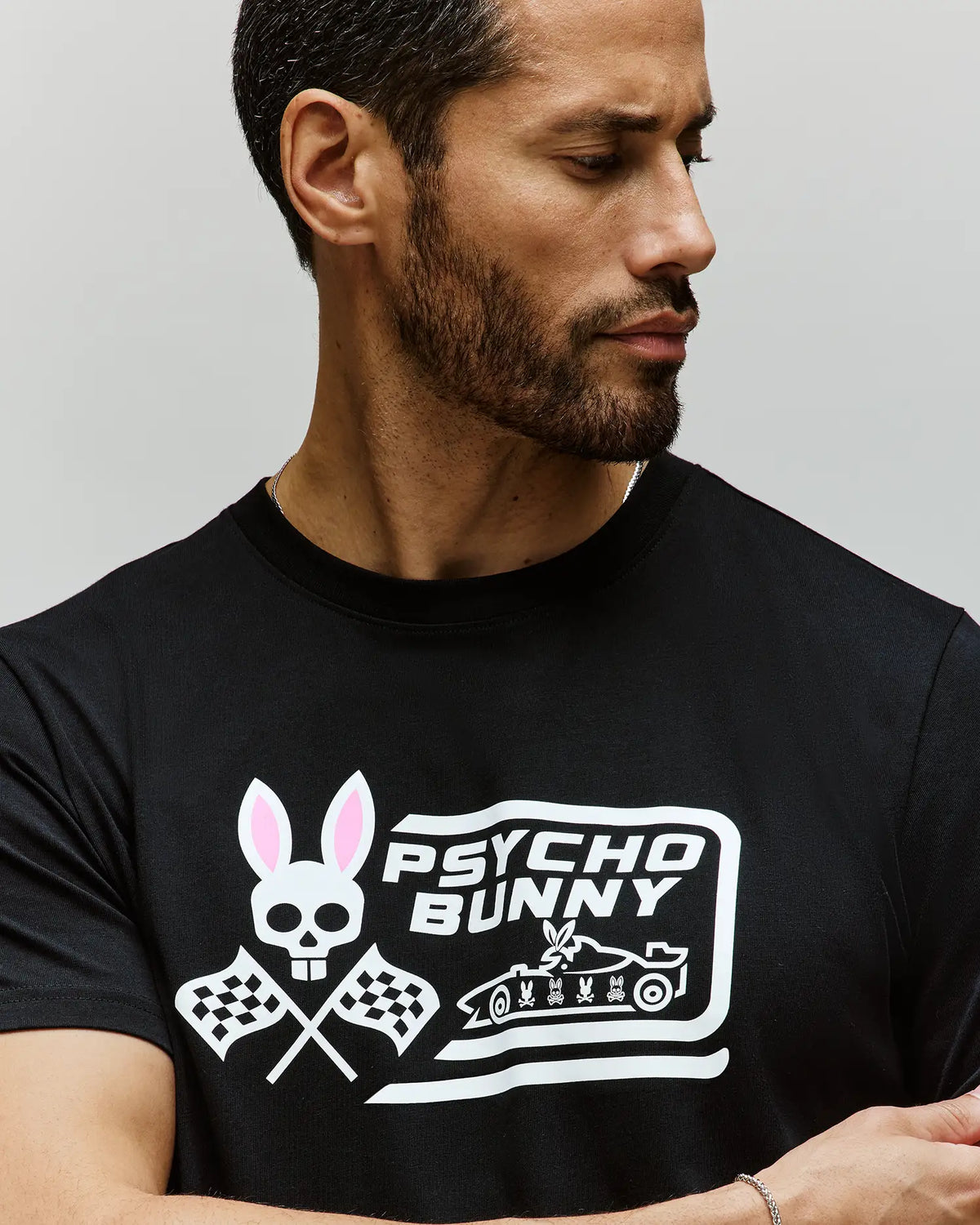 A bearded man looks left, wearing the Psycho Bunny MENS RACING GRAPHIC TEE (MT0200243) in black—its Pima cotton fabric features a skull with bunny ears, crossed racing flags, and bold "PSYCHO BUNNY" text for an edgy look.
