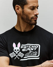 A bearded man looks left, wearing the Psycho Bunny MENS RACING GRAPHIC TEE (MT0200243) in black—its Pima cotton fabric features a skull with bunny ears, crossed racing flags, and bold "PSYCHO BUNNY" text for an edgy look.