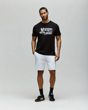 A man stands against a light gray background, wearing the Psycho Bunny MENS RACING GRAPHIC TEE (MT0200243) in black, paired with white shorts, black socks, and black Adidas sneakers. He has short dark hair, a beard, and a relaxed stance.