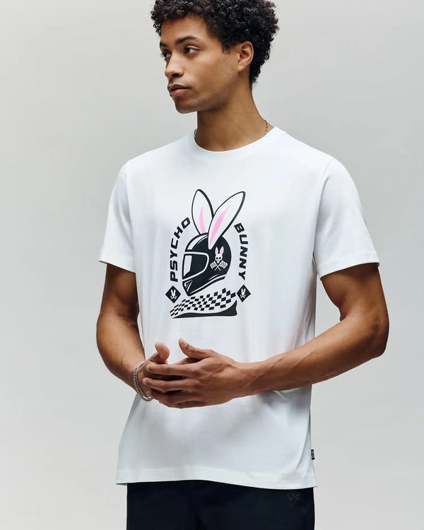 A man wears the Psycho Bunny MENS RACING BUNNY GRAPHIC TEE (MT0200245), a white Pima cotton shirt with a helmeted pink-eared bunny graphic and "Psycho Bunny" text, standing against a light gray background.