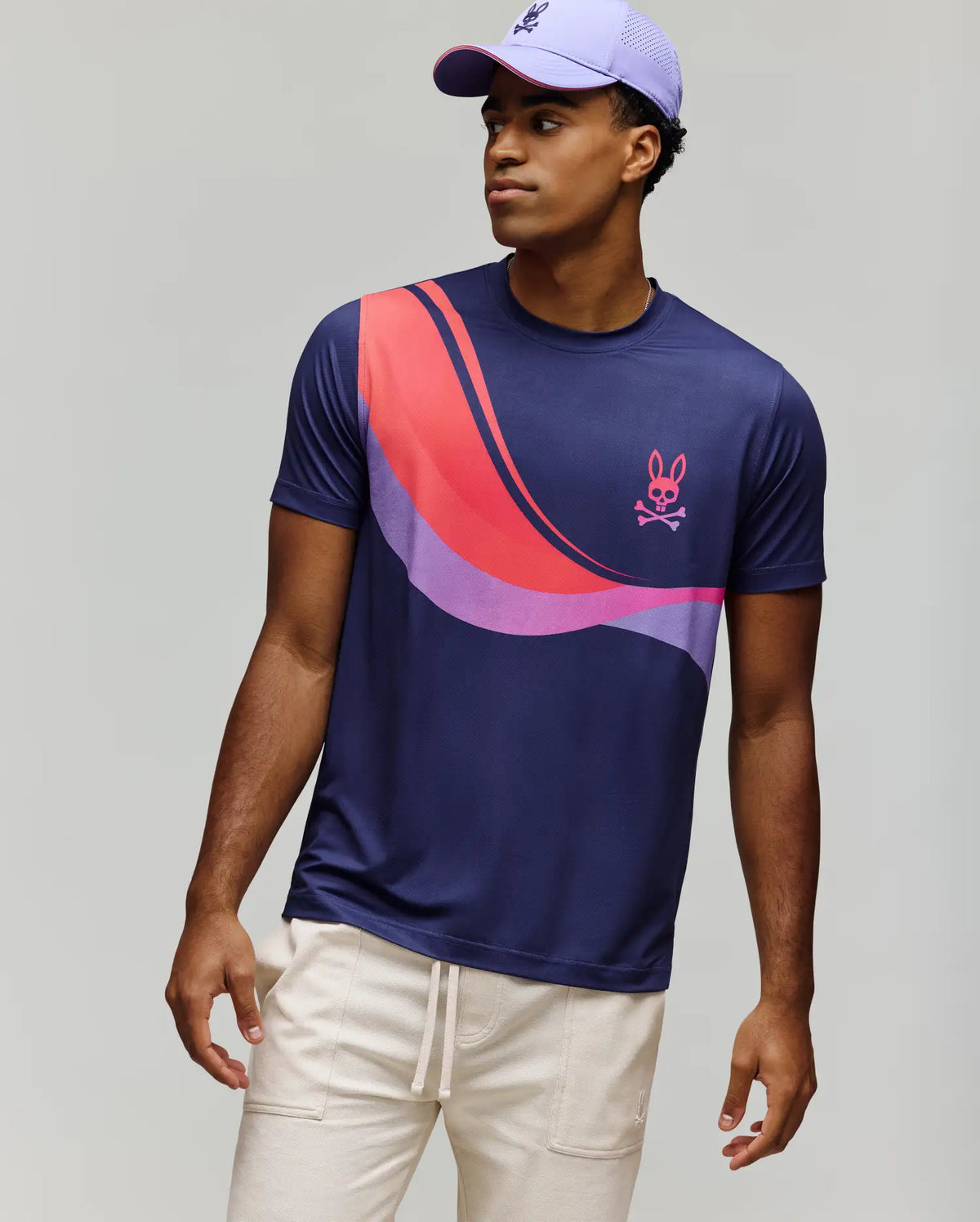A young man wears the Psycho Bunny MENS OFFICIAL US OPEN AVERY NET PERFORMANCE TEE (MT0200273), a slim fit purple and pink color-blocked tee with a bunny skull logo, paired with light beige pants and a purple baseball cap, against a gray background.