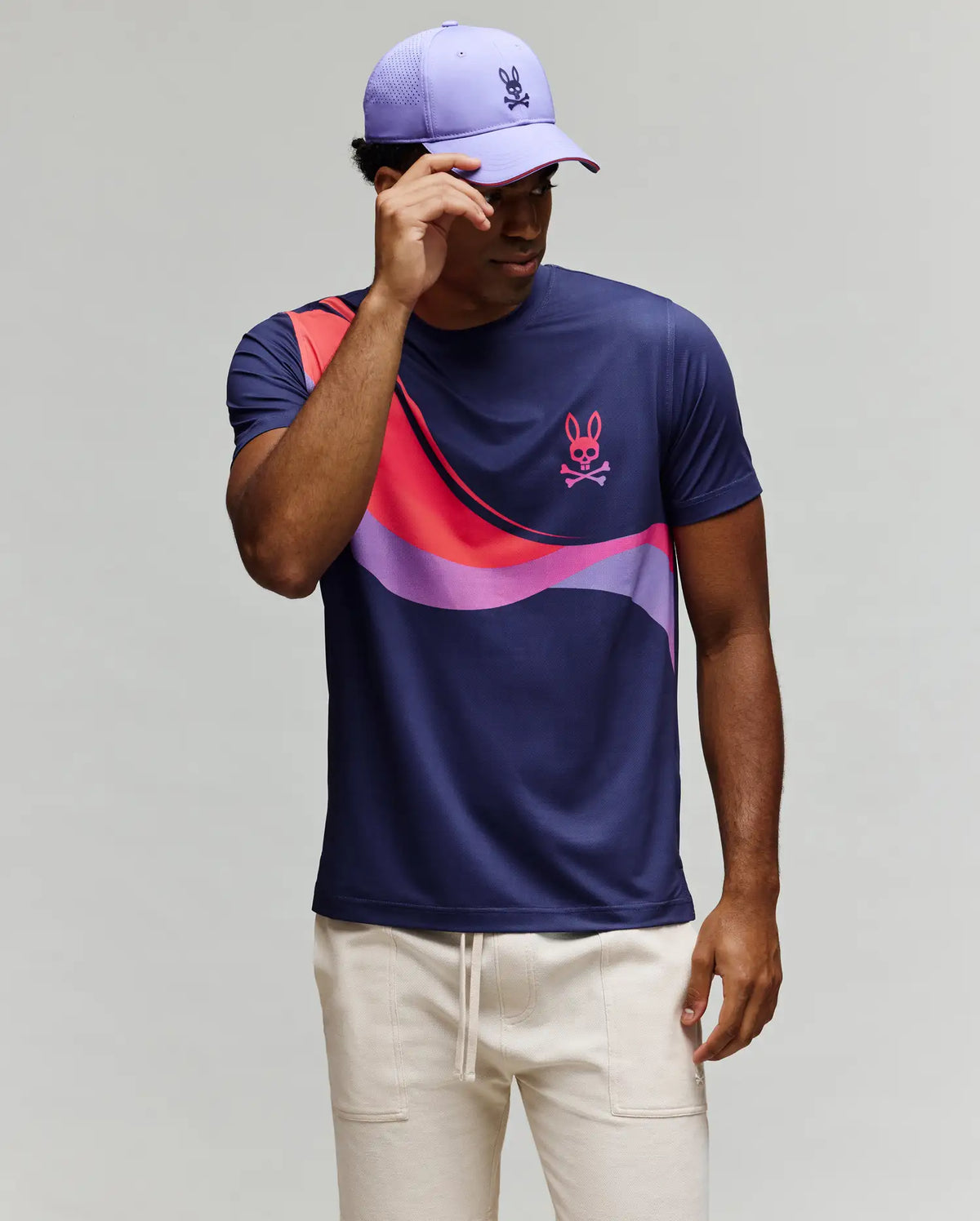 A man in light pants and a lavender cap stands against a plain background, adjusting his cap while wearing the Psycho Bunny MENS OFFICIAL US OPEN AVERY NET PERFORMANCE TEE (MT0200273) featuring navy, purple, pink, and purple wave patterns.