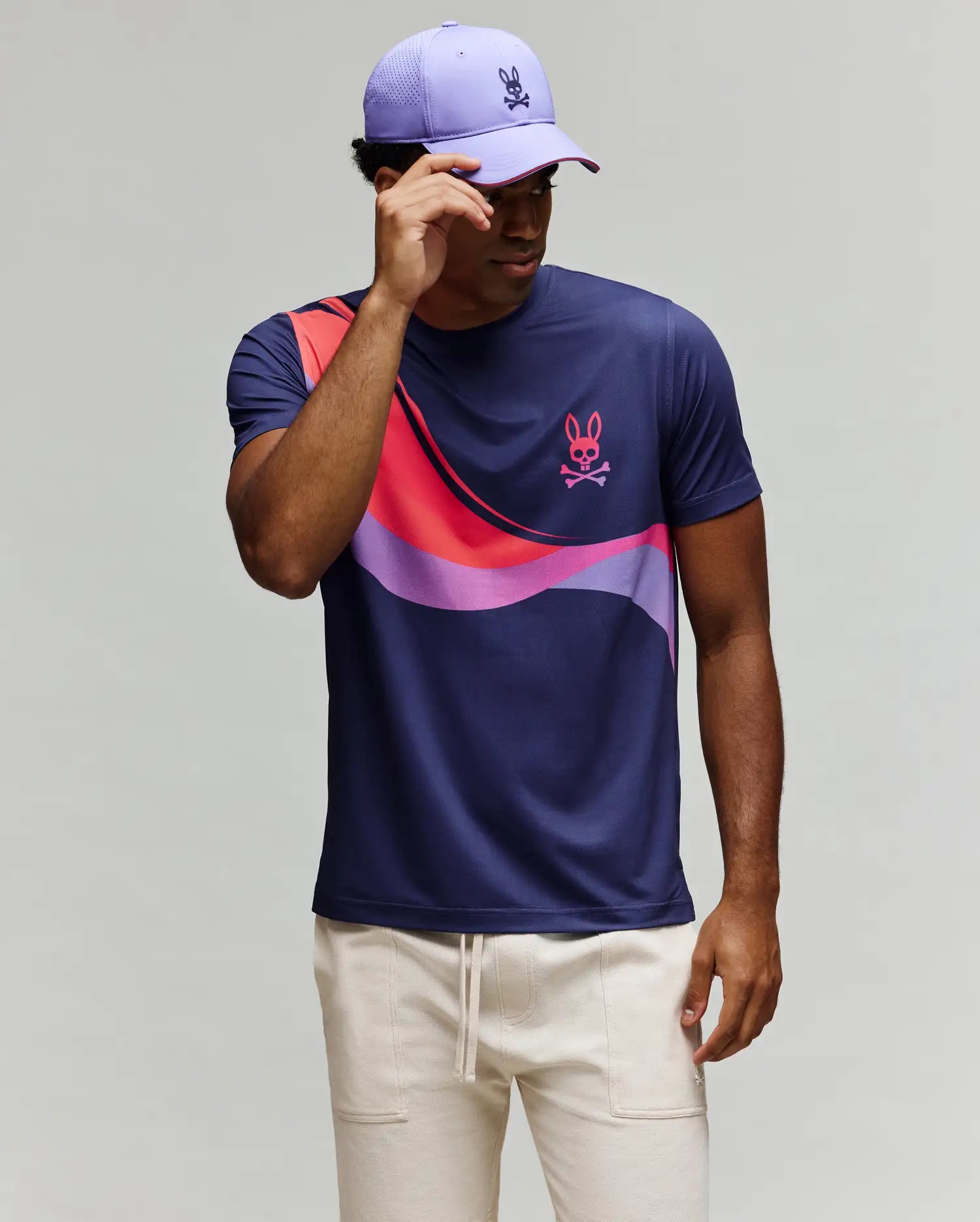 A man in light pants and a lavender cap stands against a plain background, adjusting his cap while wearing the Psycho Bunny MENS OFFICIAL US OPEN AVERY NET PERFORMANCE TEE (MT0200273) featuring navy, purple, pink, and purple wave patterns.
