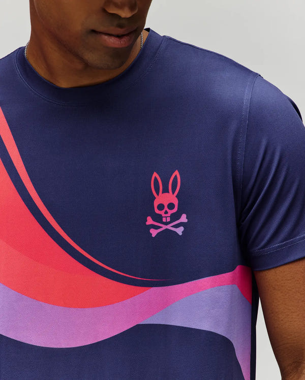 A person wears the Psycho Bunny MENS OFFICIAL US OPEN AVERY NET PERFORMANCE TEE (MT0200273), a slim fit navy blue tee with pink and purple wave patterns and a pink bunny skull & crossbones logo on the chest, showing only the lower face and upper body.