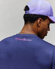 A man, turned away from the camera, wears a purple perforated cap and a navy slim fit Psycho Bunny MENS OFFICIAL US OPEN AVERY NET PERFORMANCE TEE with "PSYCHO BUNNY" and a pink-and-white bunny logo on the back collar.