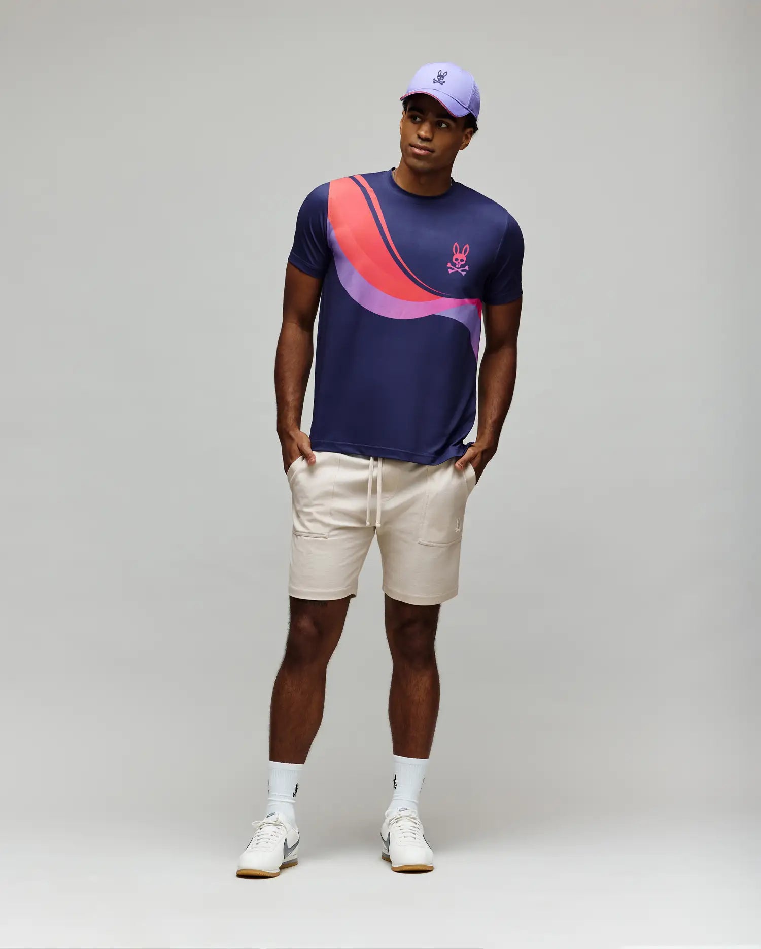 A man in a purple cap, beige drawstring shorts, white sneakers and socks wears the Psycho Bunny MENS OFFICIAL US OPEN AVERY NET PERFORMANCE TEE (MT0200273) with pink and red curved stripes, standing against a light gray background with hands in pockets.