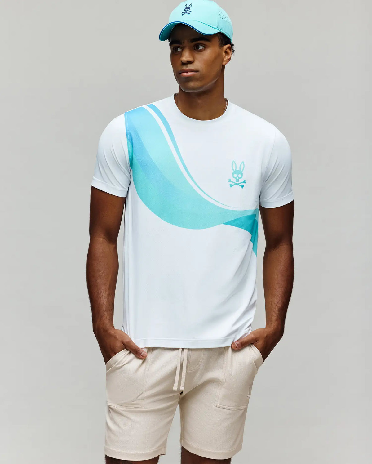 A man stands with his hands in his pockets, wearing a light blue cap, beige drawstring shorts, and the Psycho Bunny MENS OFFICIAL US OPEN AVERY NET PERFORMANCE TEE (MT0200273) against a plain background.