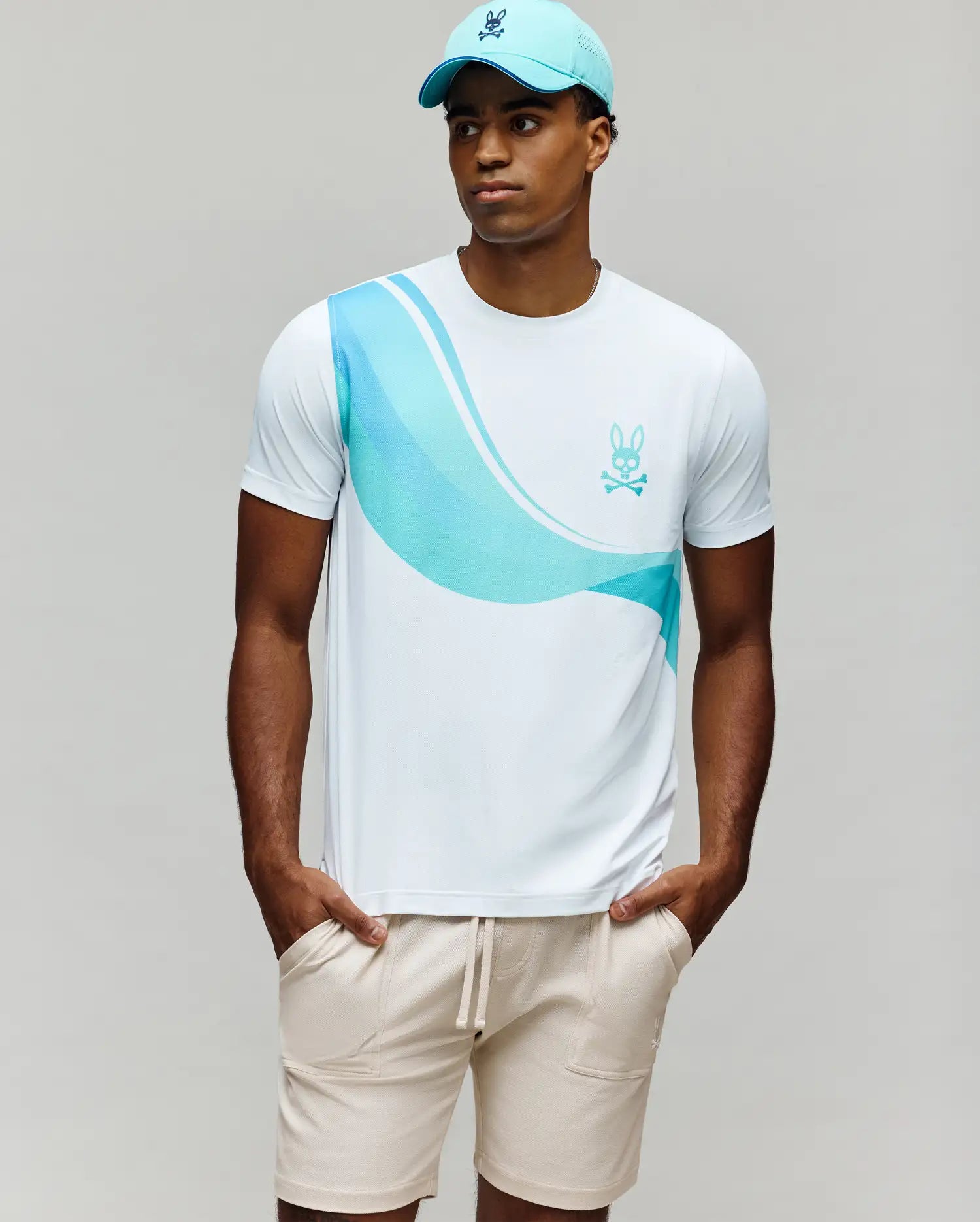 A man stands with his hands in his pockets, wearing a light blue cap, beige drawstring shorts, and the Psycho Bunny MENS OFFICIAL US OPEN AVERY NET PERFORMANCE TEE (MT0200273) against a plain background.