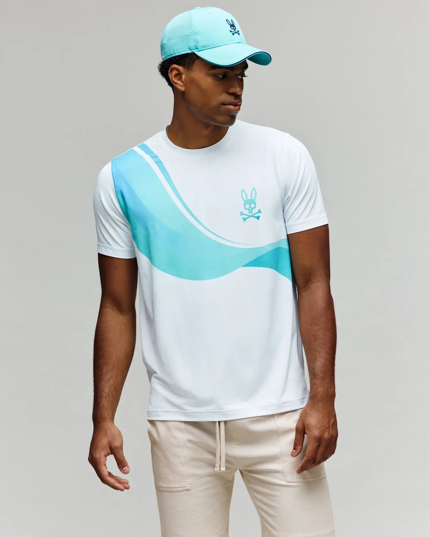 A man wearing a light blue cap, beige pants, and the Psycho Bunny MENS OFFICIAL US OPEN AVERY NET PERFORMANCE TEE (white with blue wavy stripes and bunny skull logo) stands against a neutral background.