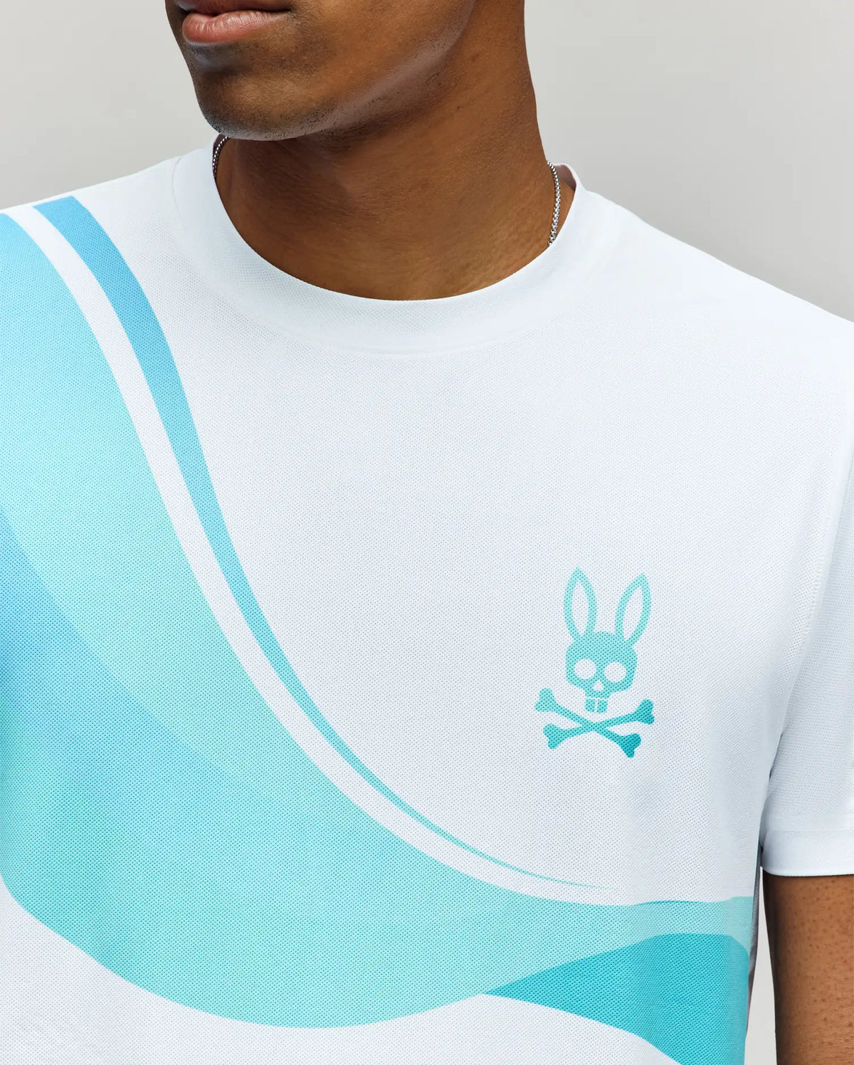 A man wears the Psycho Bunny MENS OFFICIAL US OPEN AVERY NET PERFORMANCE TEE (MT0200273), featuring white Pima cotton, light blue wave patterns, and a blue bunny skull logo. Only his lower face and upper torso are shown.