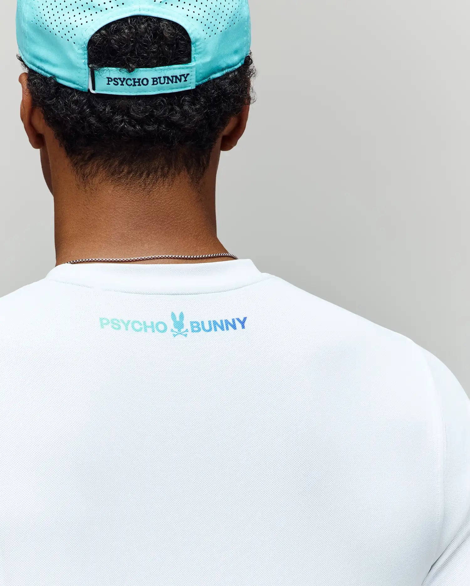 A person wearing a light blue Psycho Bunny cap and the MENS OFFICIAL US OPEN AVERY NET PERFORMANCE TEE (MT0200273), both with matching back logos, viewed from behind against a plain background.