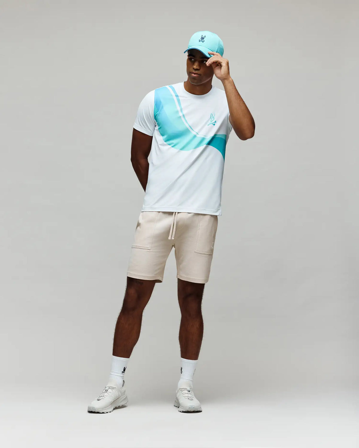 A man stands against a plain background, wearing the Psycho Bunny MENS OFFICIAL US OPEN AVERY NET PERFORMANCE TEE in white and blue, beige shorts, white sneakers and socks, plus a light blue baseball cap while holding the cap's brim.