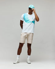 A man stands against a plain background, wearing the Psycho Bunny MENS OFFICIAL US OPEN AVERY NET PERFORMANCE TEE in white and blue, beige shorts, white sneakers and socks, plus a light blue baseball cap while holding the cap's brim.