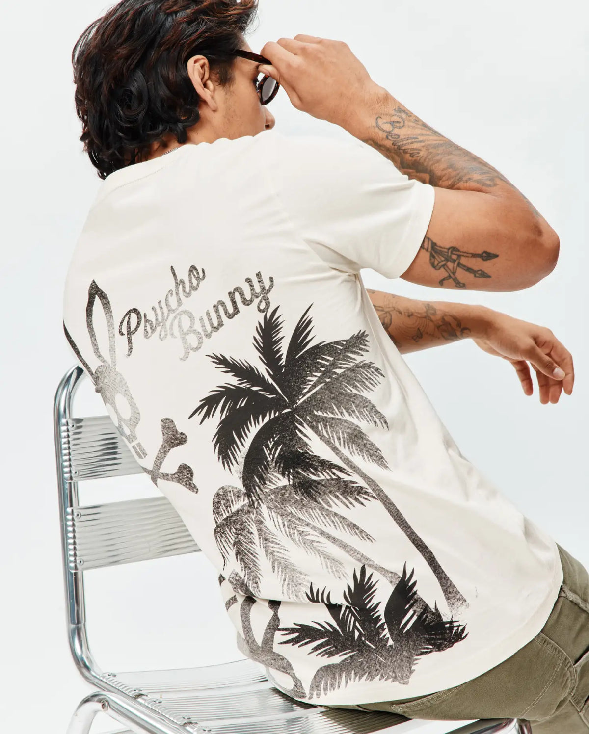 Mens Colin Back Graphic Tee, an ecru classic fit jersey knit t-shirt with a large black and grey back graphic featuring Psycho Bunny script, a bunny skull, palm trees, and crossbones. (1)