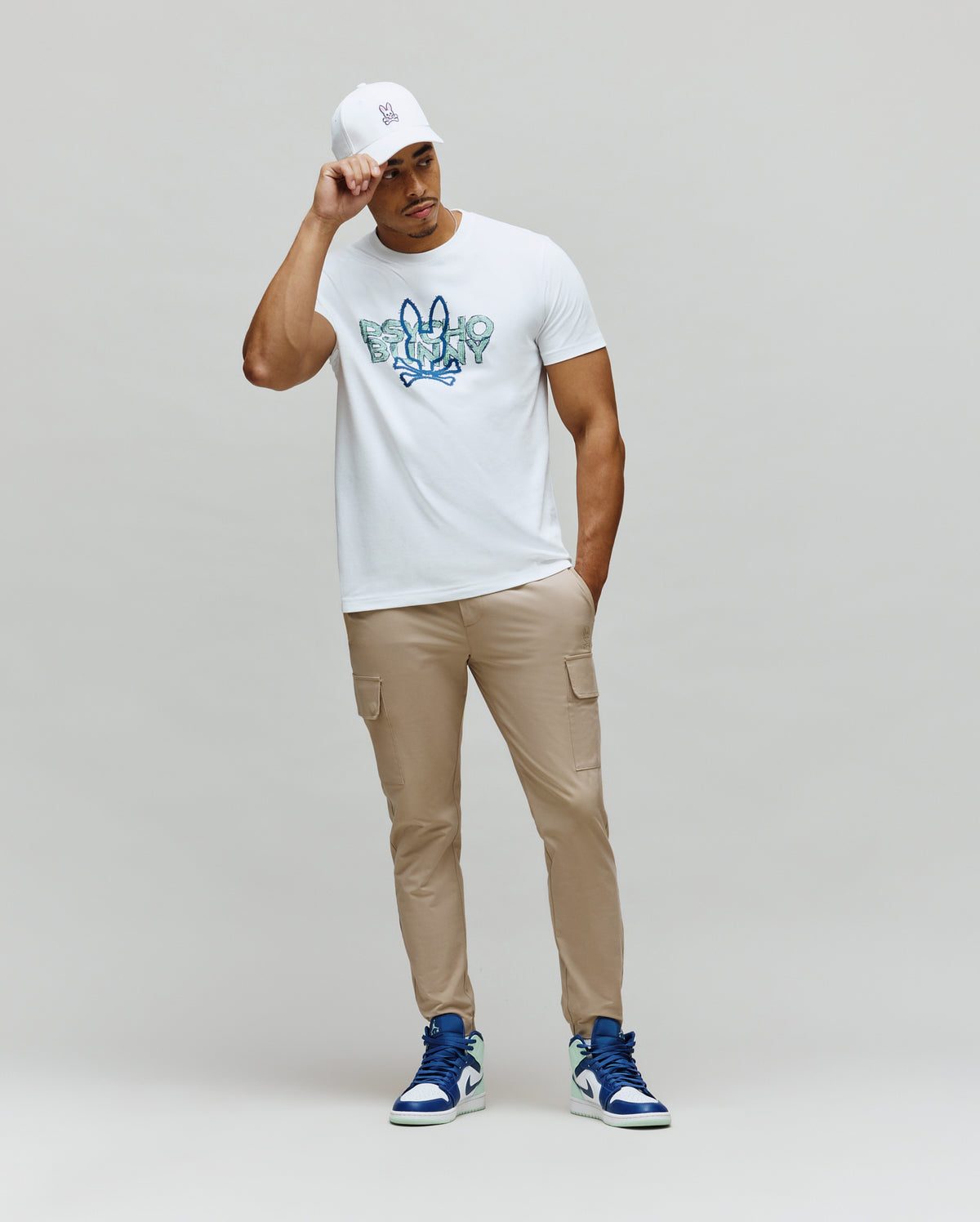 White Mens Herman Graphic Tee, classic fit, with a large blue and green textured Psycho Bunny skull and crossbones chest graphic, styled with tan slim cargo pants and a white logo cap.