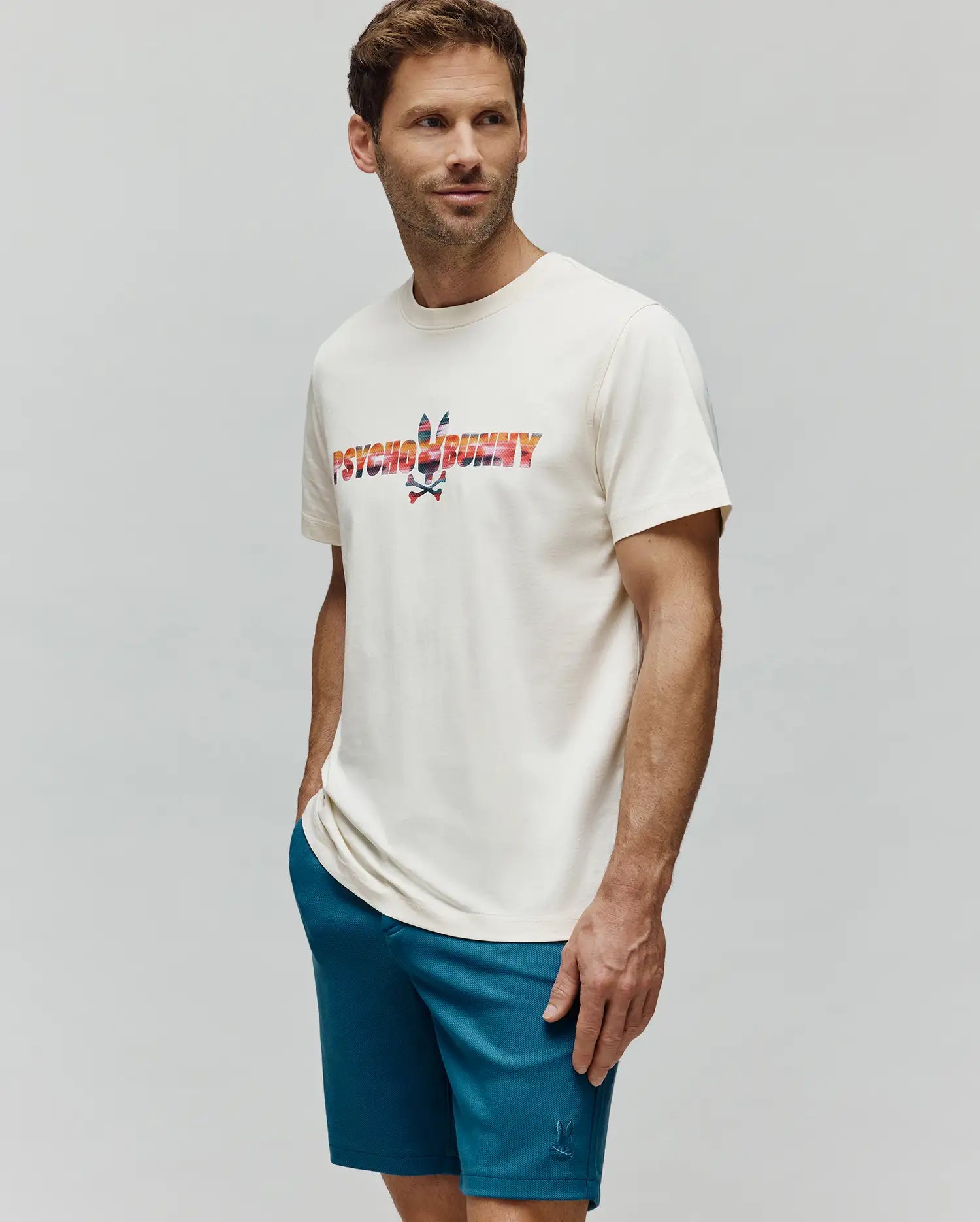 Mens Raphael Wordmark Tee in cream, classic fit, smooth jersey, with a large gradient orange-red-blue-green Psycho Bunny wordmark and skull graphic. Paired with teal pique shorts featuring a small embroidered bunny logo.