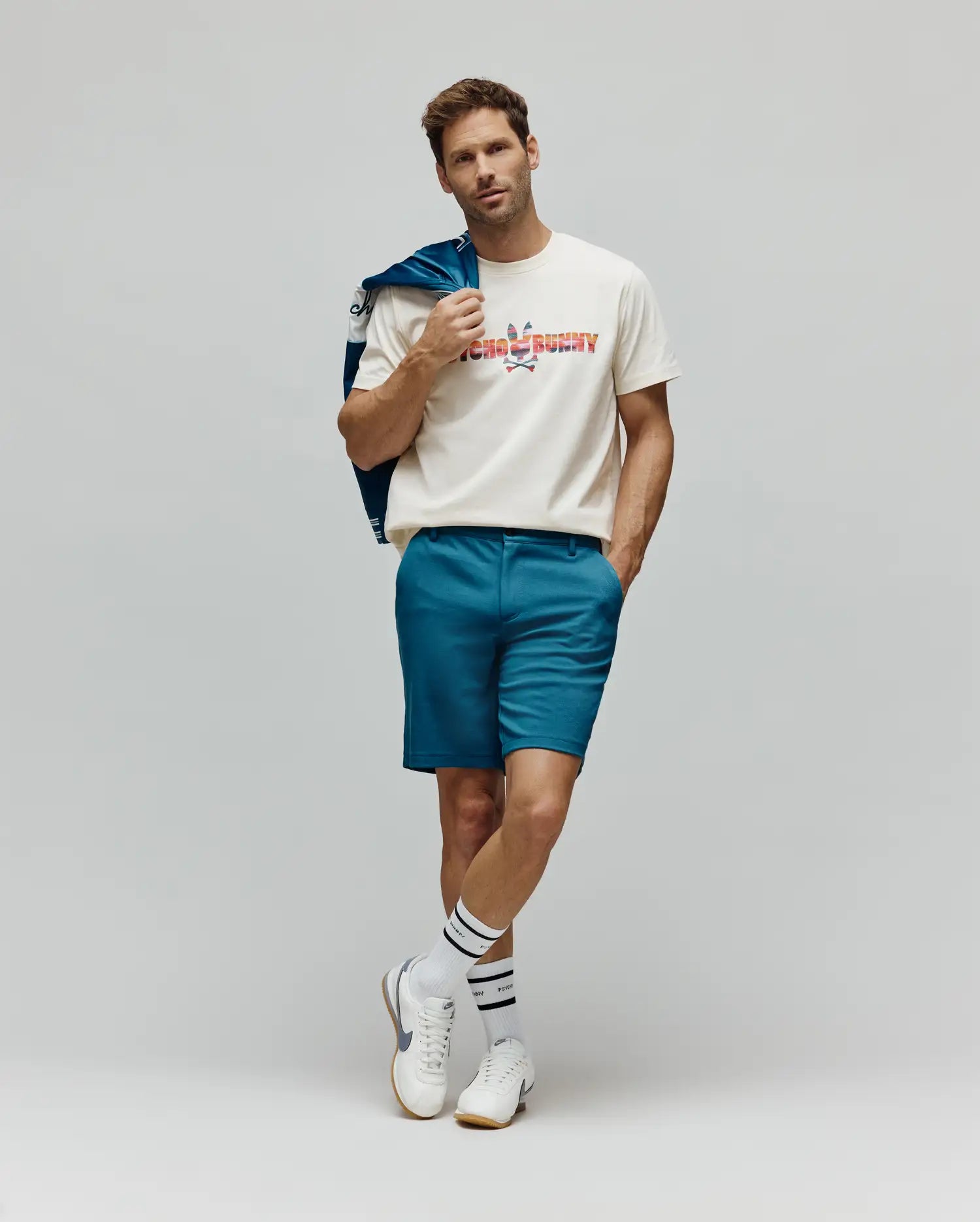 Mens Raphael Wordmark Tee, a cream jersey t-shirt featuring a large, colorful striped Psycho Bunny wordmark logo on the chest, styled with teal tailored shorts and a blue jacket.