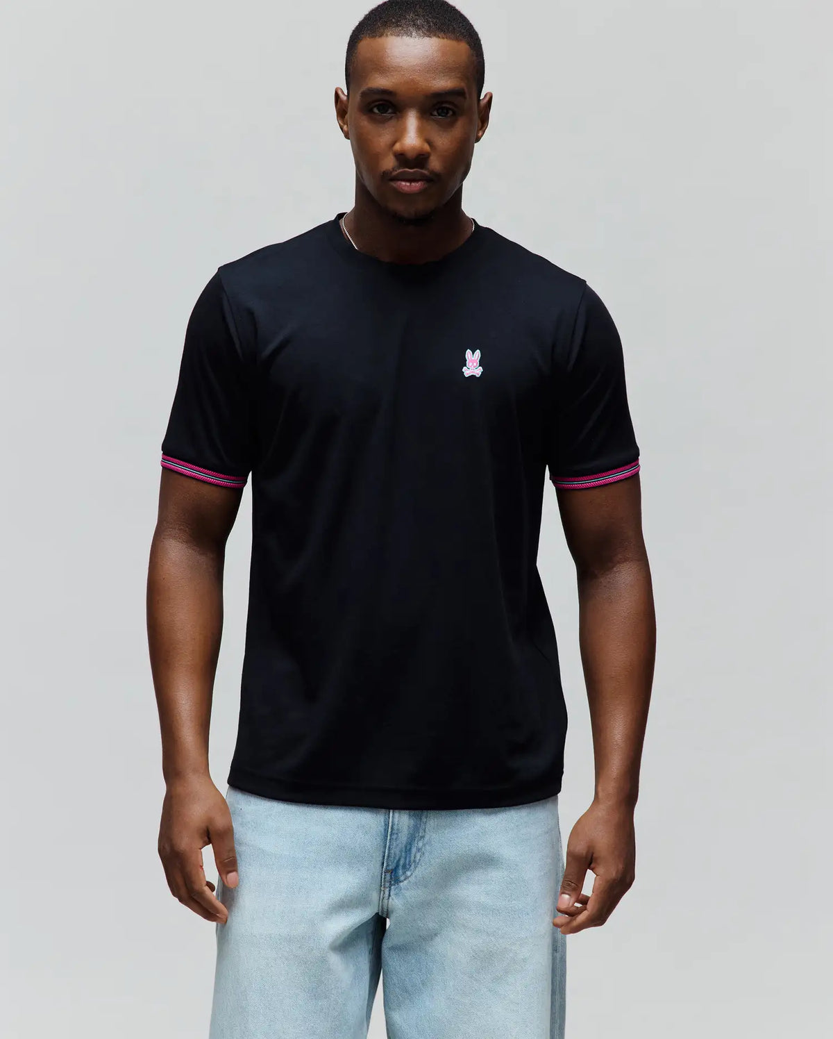 Mens Clark Performance Tee, black classic fit jersey t-shirt with small embroidered pink and white Psycho Bunny skull logo on left chest and pink and white striped sleeve cuffs.