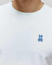 Mens Clark Performance Tee, a classic fit white t-shirt crafted from smooth jersey fabric, featuring a small, vibrant blue embroidered Psycho Bunny skull logo on the left chest.