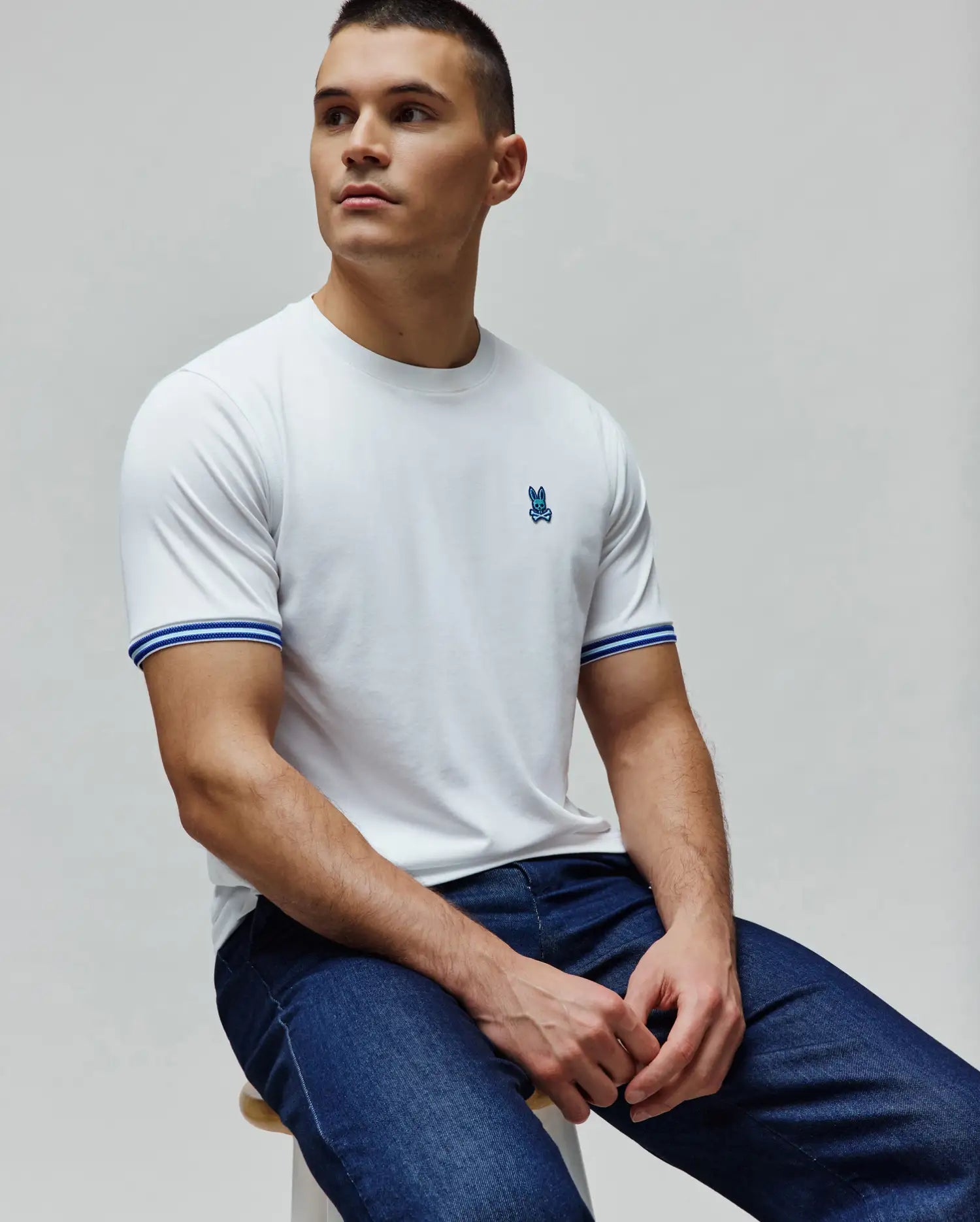 Mens Clark Performance Tee, a classic fit white performance jersey tee with a small embroidered blue Psycho Bunny skull logo on the left chest and ribbed sleeve cuffs featuring blue and grey stripes.