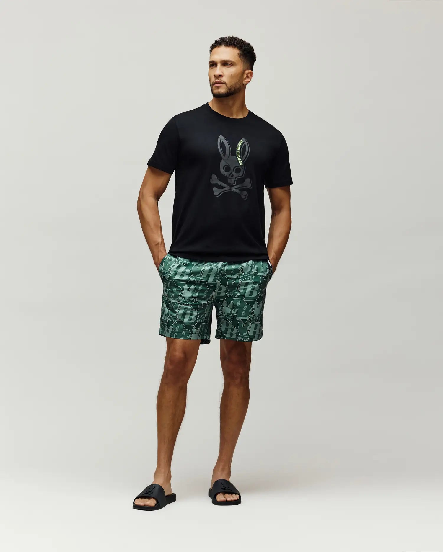 Mens Serge Embroidered Bunny Graphic Tee, a classic fit black jersey tee with a large dark grey bunny skull logo and neon green Psycho Bunny text, styled with green all-over print shorts.