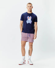 Mens John Graphic Tee, a navy blue jersey t-shirt with a large red, white, and light blue Psycho Bunny skull logo on the chest, paired with red, white, and navy striped shorts featuring a small embroidered bunny logo. (5)