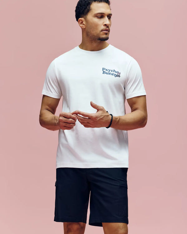 A man stands against a pink background wearing the Psycho Bunny MENS ARCHIVE WORDMARK GRAPHIC TEE (MT0200030) in white and black shorts, looking slightly to the side with his hands loosely clasped.