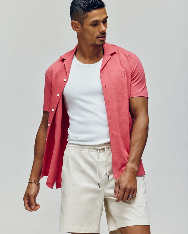 A man wearing PsychoBunny's MENS JAYME KNIT SEERSUCKER CABANA COLLAR SHIRT (MT0300002) in coral, layered over a white tank and paired with light beige drawstring shorts, stands against a plain light gray background looking to the side with one hand pocketed.