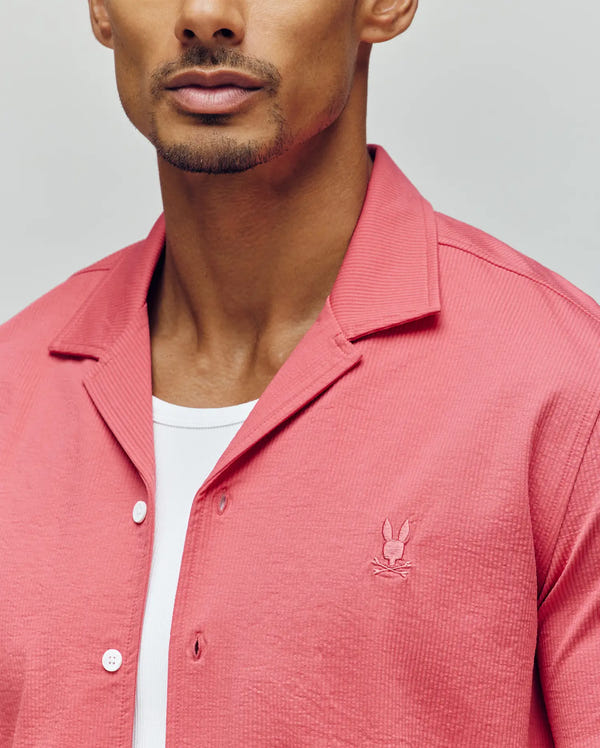 A man wears the PsychoBunny MENS JAYME KNIT SEERSUCKER CABANA COLLAR SHIRT (MT0300002), a textured pink button-up with a cabana collar over a white tee. The shirt has a small embroidered bunny logo on the chest; his face is partially visible.