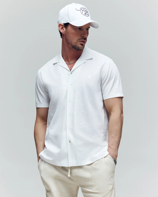 A man wearing the Psycho Bunny MENS JAYME KNIT SEERSUCKER CABANA COLLAR SHIRT (MT0300002), cream pants, and a white baseball cap stands against a light gray background with his hands in his pockets, looking to the side.